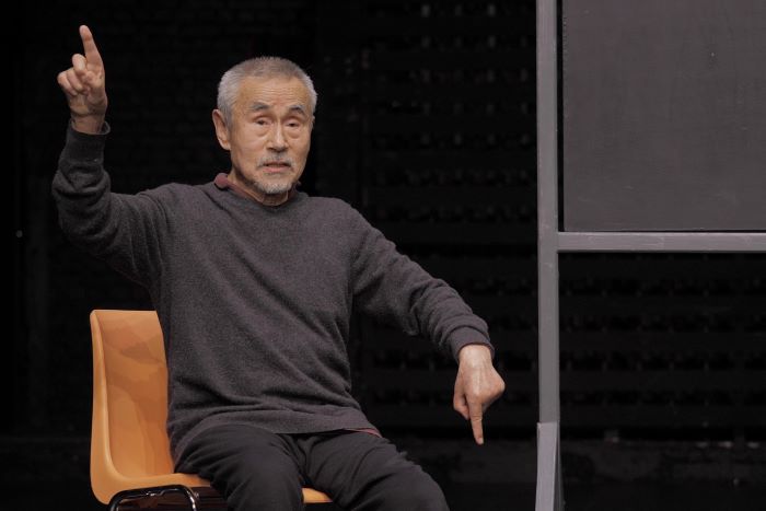 Yoshi Oida face au public tend le bras droit vers le haut et le gauche vers le bas. Ses index sont pointés.