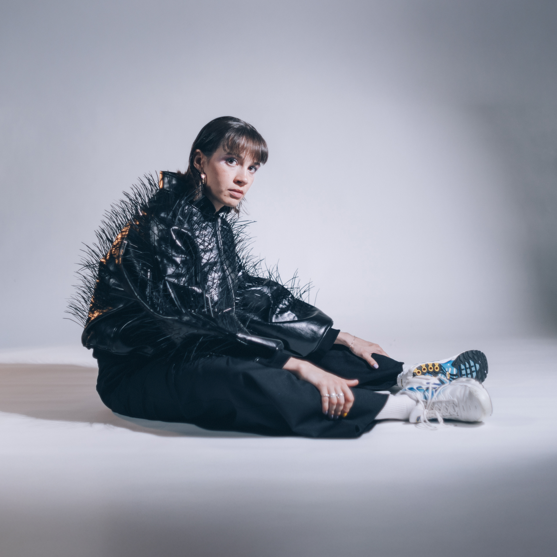 Une famme est assise par terre de profil et regarde l'objectif. Elle porte un manteau en cuir recouvert d'épines.