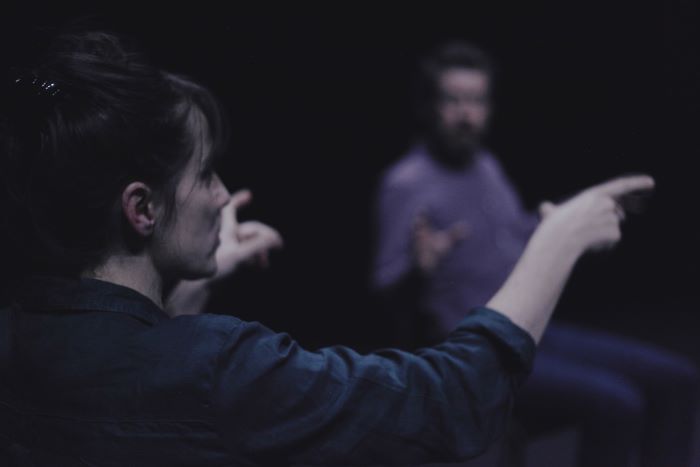 Une personne au premier plan est assise et fait des gestes pendant qu'elle parle. Au second plus, un autre personnage la regarde. Il est flou.