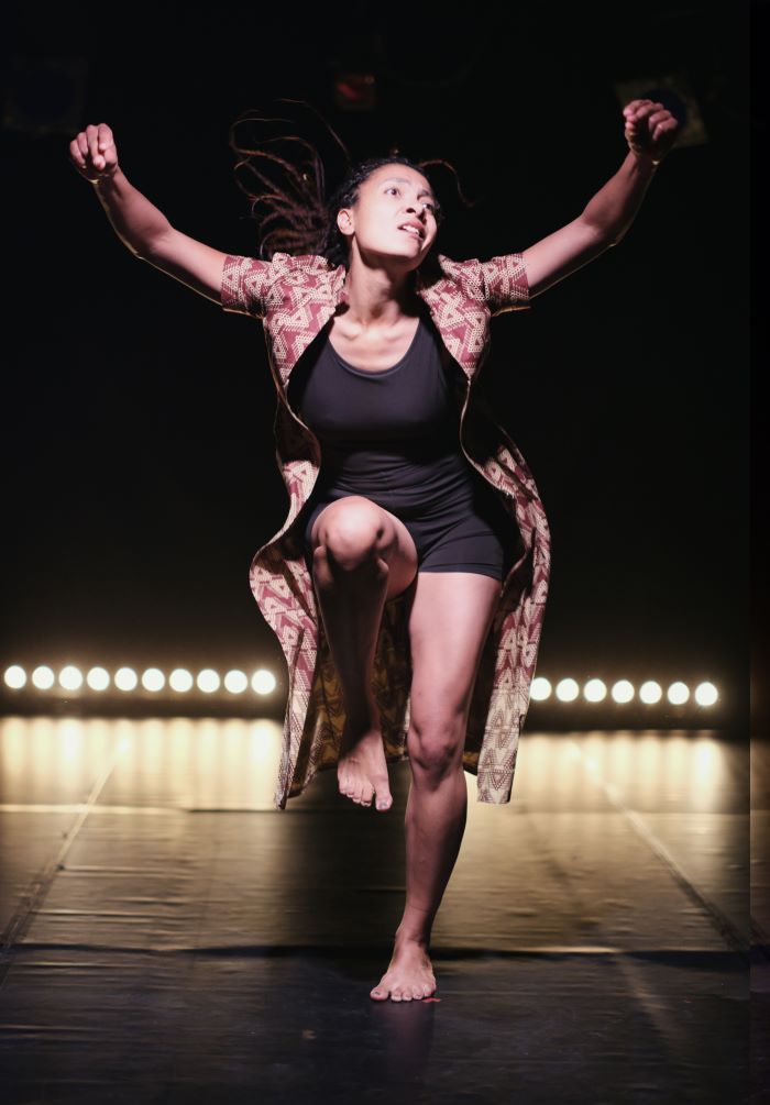 Danseuse face au public levant les deux bras vers le ciel et la jambe droite, genou plié.