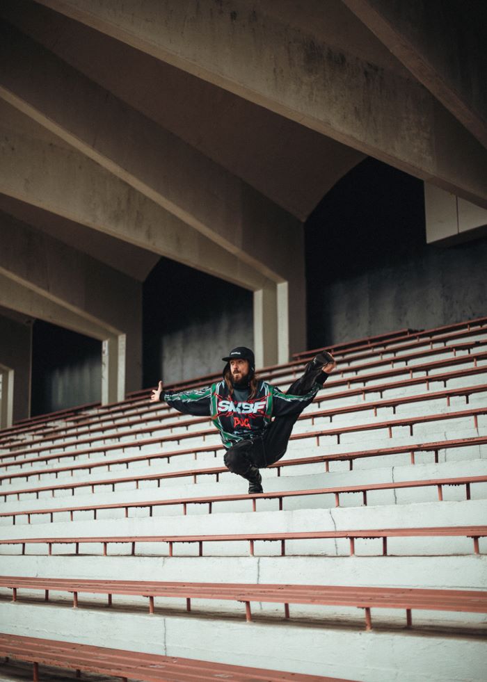 Danseur, dans un gradin, accroupi et en équilibre sur son pied droit jambe gauche tendue dans sa main. Bras droit tendu sur le côté.