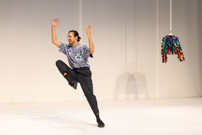 Danseur de profil en mouvement mains en l'air et jambr droite repliée. En fonds de scène une veste suspendue à un cintre.
