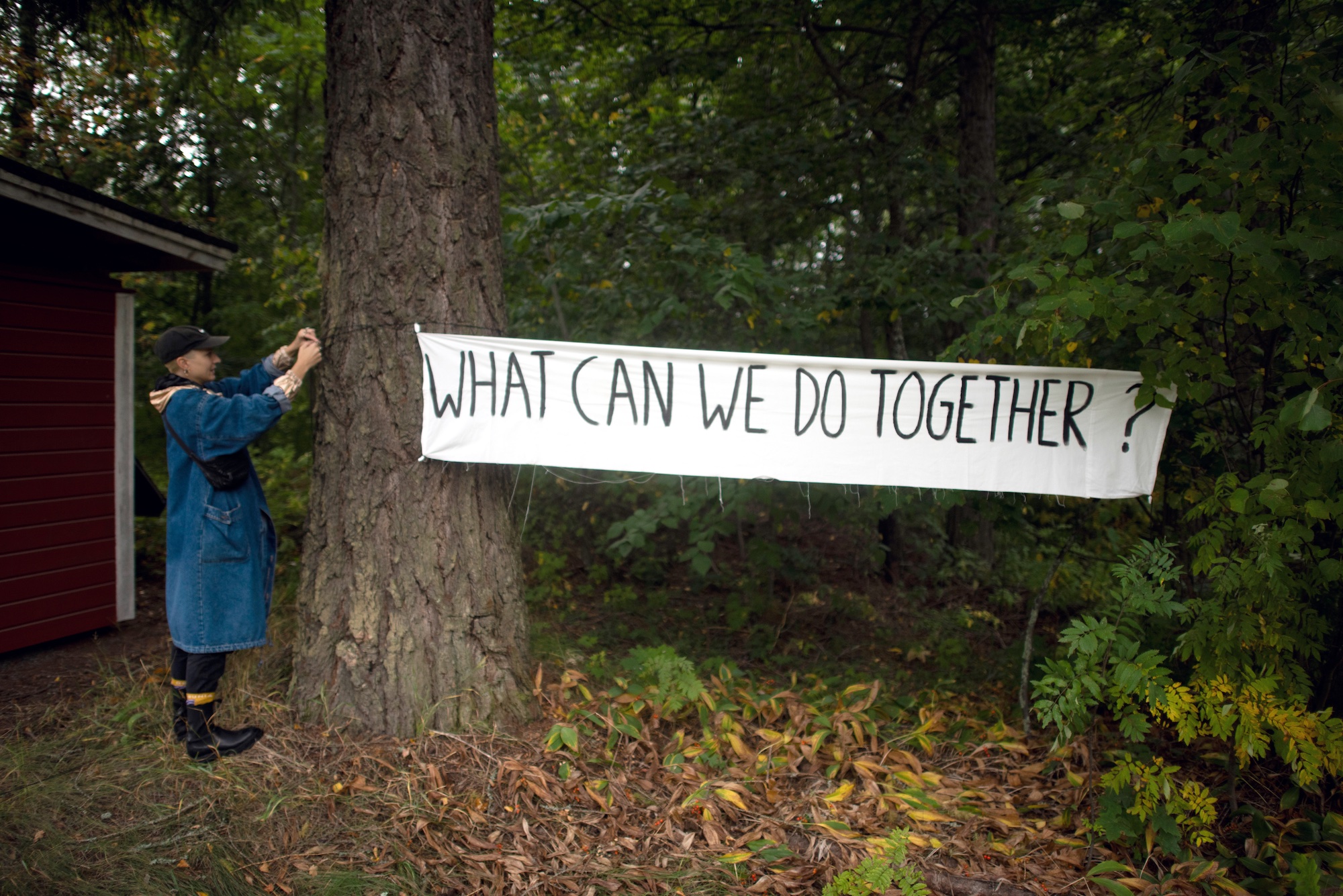 Une personne prend en photo une banderole attachée entre deux arbres où il est écrit : "What can we do together?"